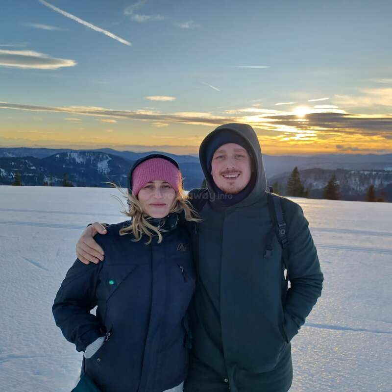 Ein lächelndes Paar, warm angezogen, steht zusammen in einer verschneiten Landschaft bei Sonnenuntergang. Im Hintergrund sieht man Berge und Bäume unter einem bunten Himmel.