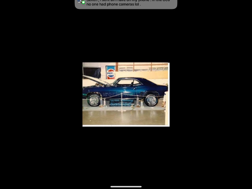 Um clássico muscle car azul está exposto dentro de casa, atrás de uma barreira de corda, com um letreiro da Pepsi ao fundo. O capô do carro está aberto para visualização.