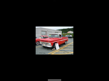 Uma caminhonete vintage vermelha brilhante está estacionada ao ar livre em uma exposição de carros. Uma placa informativa está em frente. Carros clássicos e prédios estão ao fundo.