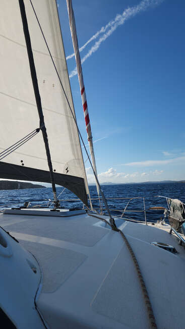 A sailboat glides on deep blue water under clear skies. The sail is up, wind fills it; distant land and white clouds complete the serene scene.