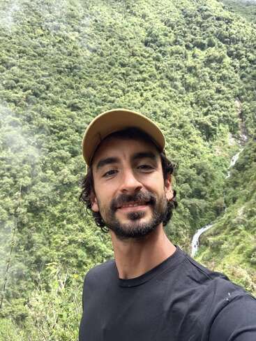 Um homem usando um boné amarelo e uma camisa preta sorri para uma selfie. Atrás dele, há uma montanha verdejante e arborizada e uma pequena cachoeira.