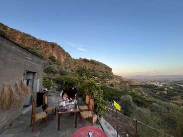 Uma pessoa arruma a mesa num terraço com vista para colinas verdejantes e um vale ao longe, sob um céu azul claro ao pôr do sol. Um cenário tranquilo.