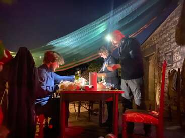 Três pessoas estão reunidas ao redor de uma mesa ao ar livre à noite, iluminadas por lanternas de cabeça e pela luz ambiente. Comida, bebidas e utensílios de cozinha estão espalhados pelo local, criando um ambiente animado e rústico.