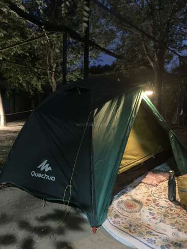 Uma barraca verde da marca Quechua está montada ao ar livre ao anoitecer, iluminada por dentro. Um tapete colorido, uma garrafa e uma bolsa estão colocados na entrada da barraca.