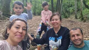 Una familia de cinco miembros disfruta de un picnic en un frondoso bosque verde. Dos niños permanecen detrás, uno de ellos saludando, mientras tres adultos sonríen a la cámara.