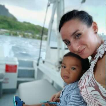 The image depicts a woman and a child sitting on a boat, with the child holding a blue object, set against a scenic backdrop of water and greenery.