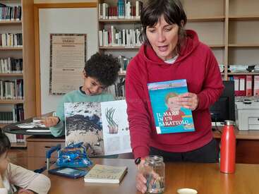 A woman in a red hoodie holds a book titled "Il mare in un barattolo" while teaching children in a library, with books and a jar on the table.