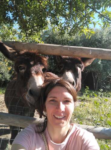 Una mujer sonriente se hace un selfi al aire libre con dos simpáticos burros marrones detrás de una valla de madera. La escena es soleada, rodeada de vegetación y árboles.