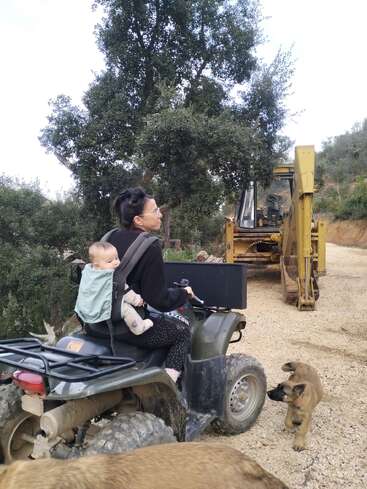 A woman carries a baby on her back while riding an ATV on a gravel road. A puppy walks beside them and construction equipment is visible ahead.