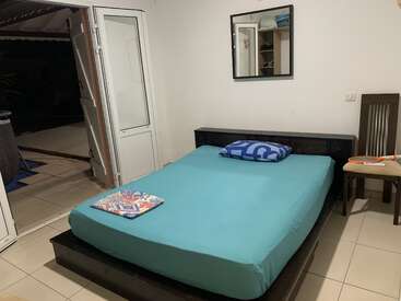 A tidy, minimalist bedroom with a turquoise sheeted bed, a blue pillow, and a colorful towel. Open door to a patio, chair, mirror, and white walls.