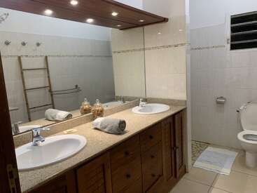 This is a modern bathroom with a large mirror, double sinks, wooden cabinets, decorative jars, folded towels, a white toilet, and bright built-in lighting.