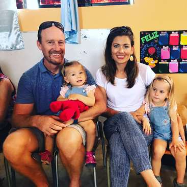 A imagem mostra uma família de quatro pessoas sentadas juntas em uma sala de aula, com o pai segurando um bebê e a mãe e duas filhas pequenas sentadas ao lado dele.