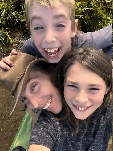 Three people, two children and one adult, are smiling brightly while taking a close-up selfie outdoors. Green grass and sunlight create a joyful, cheerful atmosphere.