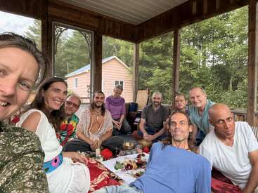 A group of people sit together on a covered porch, smiling and relaxed. The setting appears peaceful, with trees and a small house in the background.