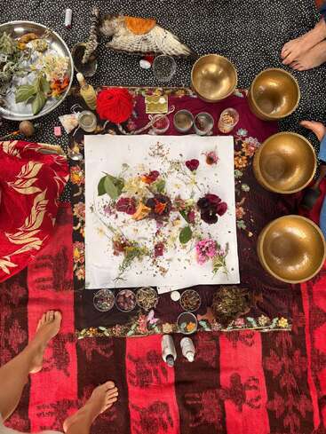 This image shows a ceremonial or spiritual setup with bowls, herbs, flowers, feathers, candles, red yarn, and bare feet on colorful, patterned cloths. Ritualistic, vibrant scene.
