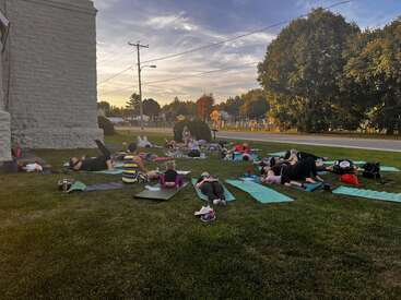 A group of people practice yoga outdoors on a grassy lawn at sunset, lying on mats near a building, with trees and street in the background.