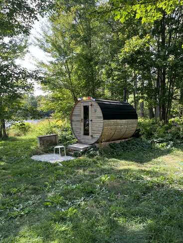 A small, barrel-shaped wooden sauna sits surrounded by lush green trees and grass. Nearby, a white stool and concrete slab create a peaceful, natural retreat.