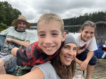 A happy family enjoys time together outdoors by a lake. They smile for a selfie on a wooden dock, surrounded by trees and cloudy skies.