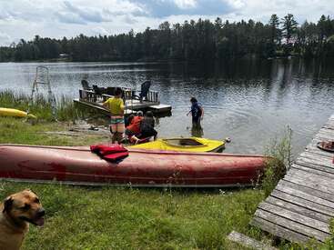 Children play near the shore of a calm lake, with kayaks, a dock, and a dog in the foreground. Forest surrounds the peaceful, summer scene.