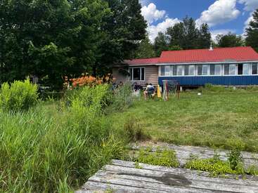A cozy house with a red roof sits amid lush greenery. Wildflowers bloom. People relax on the lawn with outdoor games, enjoying a sunny summer day.