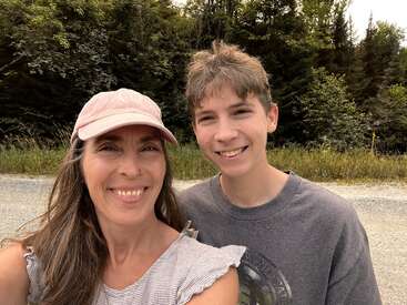 A woman and a teenage boy smile for a selfie outdoors, standing on a gravel road with green trees in the background under a sunny sky.