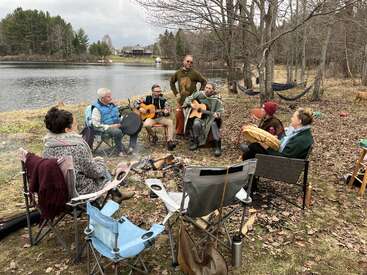 A group of people sit around a campfire by a lake, playing instruments, singing, and enjoying nature in a relaxed, friendly outdoor gathering.