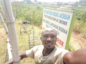 A man takes a selfie outdoors near a restoration project sign. Another person works with wooden structures in the background. Lush greenery surrounds the area.