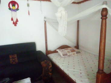 The image shows a cozy bedroom with a wooden four-poster bed, a mosquito net, patterned bedsheet, pillows, a black sofa, and wall hanging décor.