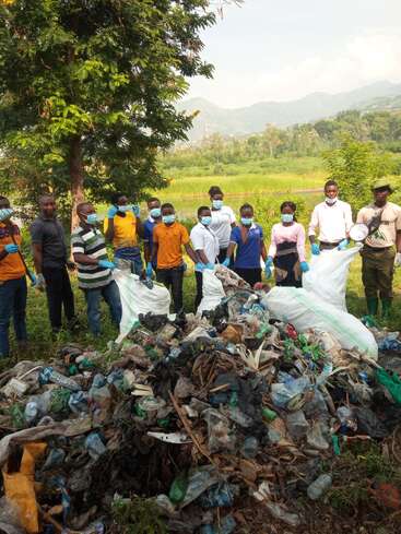 A group of people wearing masks and gloves stand outdoors, having collected a large pile of plastic bottles and waste for environmental cleanup in nature.