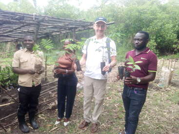 Four people stand outdoors, each holding a young tree seedling. Behind them is a wooden structure and lush greenery, indicating a tree planting or environmental activity.