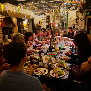 Eine lebhafte Gruppe von Menschen sitzt an einer langen Tafel, umgeben von funkelnden Lichtern, Dekorationen, Essen, Getränken und warmen, fröhlichen Gesprächen.
