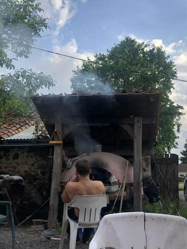 Ein Mann ohne Hemd sitzt auf einem weißen Plastikstuhl und schaut auf einen rustikalen Holzofen im Freien. Rauch steigt auf, umgeben von Bäumen, Stühlen und einem Steinbau. Entspannte Atmosphäre.