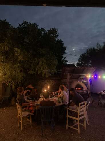 Eine Gruppe von Freunden sitzt nachts draußen an einem Tisch, beleuchtet von Kerzen und Lichterketten, und neben ihnen steht ein Pizzaofen, der gemütlich leuchtet.