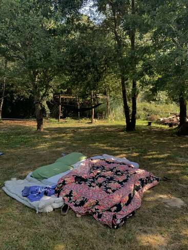 Ein gemütliches Bett im Freien mit bunten Decken und Kissen steht unter schattigen Bäumen auf einer ruhigen Wiese, mit einer Hängematte und Holzstühlen im Hintergrund.