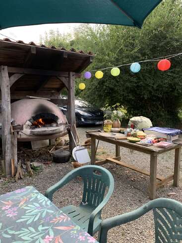 Eine Szene im Freien mit einem Holzofen, einem Tisch mit Lebensmitteln, grünen Stühlen, einer geblümten Tischdecke, bunten Laternen, Bäumen und einem Auto, die eine festliche Atmosphäre schaffen.