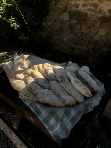 Ein Stapel frisch gebackener rustikaler Brote liegt draußen auf einem karierten Tuch, und die Sonne wirft Schatten neben einer alten Steinmauer und viel Grün.