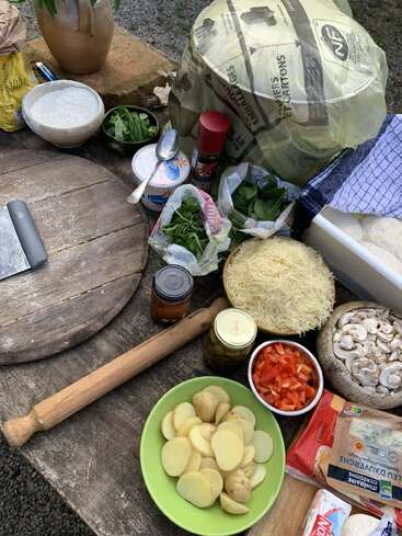 Auf dem Bild sieht man einen rustikalen Tisch mit verschiedenen Zutaten für Pizza: geschnittene Kartoffeln, Käse, Pilze, Paprika, Gemüse, Mehl, Joghurt und Küchenutensilien, alles bereit zum Kochen.