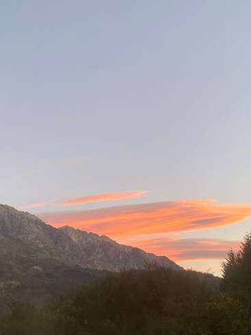 Un sereno paisaje montañoso al atardecer, con nubes de color naranja intenso que atraviesan un cielo despejado, una exuberante vegetación debajo y una suave luz vespertina que realza la belleza natural.