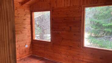 Esta imagen muestra el interior de una pequeña cabaña de madera con paredes de madera natural, dos grandes ventanas y vistas a los árboles y la vegetación del exterior. Ambiente acogedor.