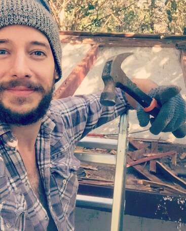 A bearded man in a plaid shirt and knit beanie smiles, holding a hammer and wearing gloves, standing outdoors on a construction project amid wooden beams.