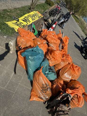 Varias bolsas de basura naranjas y verdes llenas de residuos recogidos están apiladas en una acera. Cerca de allí se ven una pancarta amarilla, bicicletas, escombros y una persona.