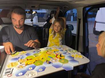 Un hombre y dos niños están jugando juntos a un juego de mesa dentro de una furgoneta, sentados alrededor de una mesa; parecen estar muy entretenidos y felices en ese ambiente acogedor.