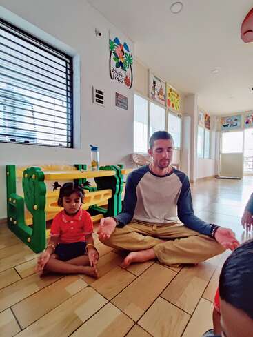 Ein Mann und ein junges Mädchen sitzen im Schneidersitz auf dem Boden und meditieren gemeinsam in einem farbenfrohen Klassenzimmer, das mit Kinderkunstwerken, Spielzeug und hellem Tageslicht gefüllt ist.