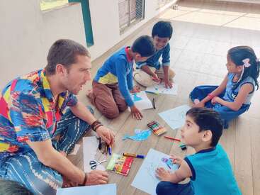 Eine Lehrerin und fünf kleine Kinder sitzen auf dem Boden und malen gemeinsam. Die Kinder lächeln und benutzen Buntstifte und Papier.