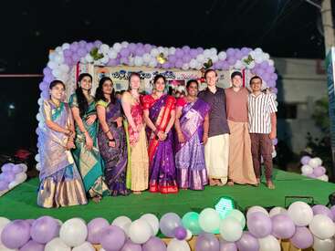 Eine Gruppe von Menschen in traditioneller indischer Kleidung posiert auf einer dekorierten Bühne mit Luftballons. Sie lächeln und feiern gemeinsam ein Ereignis in der Nacht.