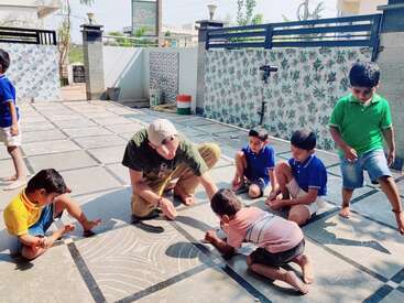 Eine Gruppe von Kindern und ein Erwachsener malen mit Kreide auf einem gefliesten Boden im Freien und genießen das gemeinsame kreative Spiel bei strahlendem Sonnenschein.