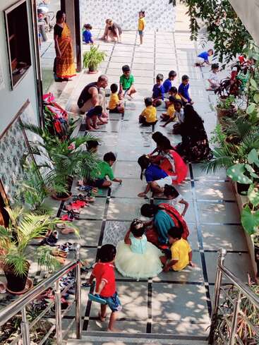 Kinder sitzen auf dem gefliesten Boden und malen mit Kreide, beaufsichtigt von Erwachsenen. Die Schuhe sind an der Wand aufgereiht. Pflanzen sorgen für Grün und eine lebendige Atmosphäre.