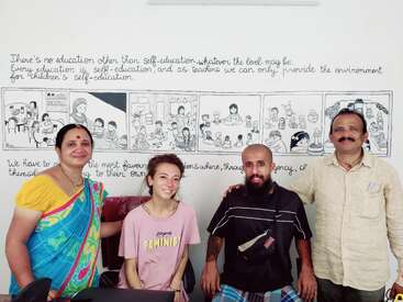 Vier Erwachsene, drei Männer und eine Frau, posieren lächelnd vor einem pädagogischen Wandbild. Das Wandbild zeigt Illustrationen und inspirierende Zitate über Selbsterziehung und Unterricht.