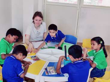 Eine Lehrerin hilft fünf kleinen Kindern an einem runden Tisch beim Malen. Die Kinder arbeiten konzentriert mit Farbstiften, Buntstiften und Markern.