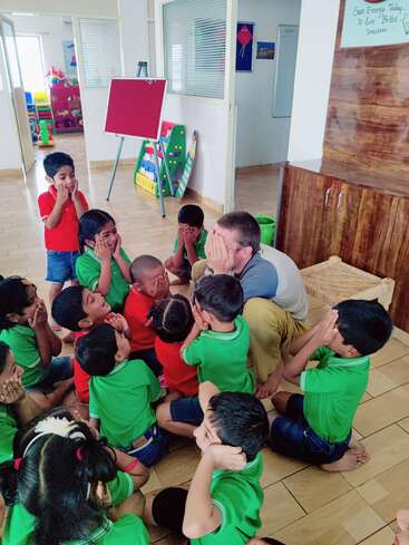 Eine Gruppe kleiner Kinder und ein männlicher Lehrer sitzen in einem Klassenzimmer auf dem Boden, bedecken spielerisch ihre Gesichter und nehmen an einer spannenden Gruppenaktivität teil.